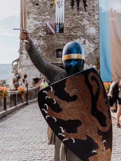 Ein Ritter in einer Rüstung steht auf einer Brücke vor einer Burg. Er hält ein Schwert in der Hand und zeigt ein Wappen auf seinem Schild. | © TV Hochsteiermark, nicoleseiser.at