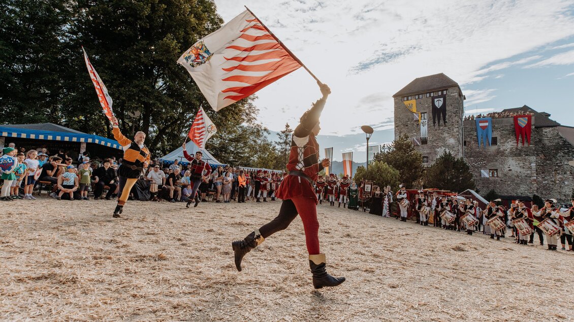 Eine lebendige Feier mit Leuten in historischer Kleidung, die Fahnen schwenken. Im Hintergrund ist eine Burg und eine große Menge Zuschauer zu sehen. | © TV Hochsteiermark, nicoleseiser.at