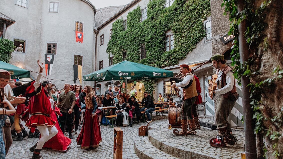 Eine lebhafte Veranstaltung mit Tänzern und Musikern in einem historischen Innenhof. Die Zuschauer genießen die Unterhaltung unter grünen Markisen. | © TV Hochsteiermark, nicoleseiser.at