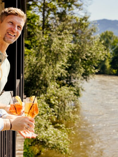 Ein Paar steht auf einem Balkon und hält Cocktails in der Hand. Im Hintergrund fließt ein Fluss, umgeben von Bäumen und Bergen.
