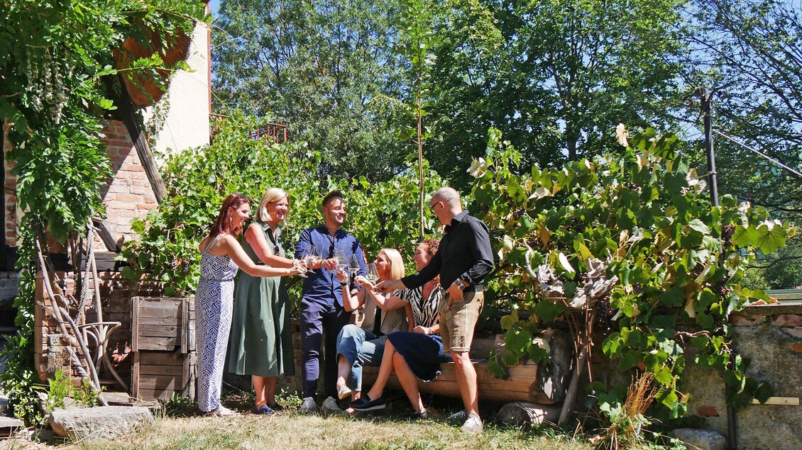 Eine Gruppe von sechs Personen steht in einem Garten und feiert gemeinsam. Sie lächeln, während sie Getränke anstoßen, umgeben von viel Grün. | © Stadt Bruck - L.Schneller