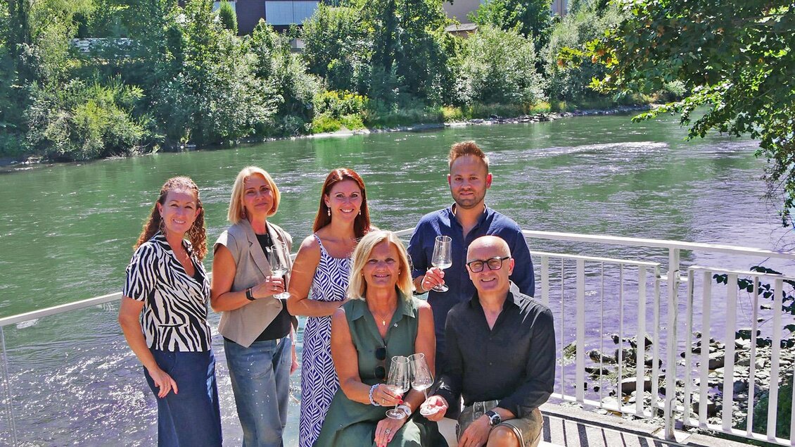 Eine Gruppe von sechs Personen posiert am Ufer eines Flusses vor einer grünen Landschaft. Im Hintergrund sind Bäume und Gebäude sichtbar. | © Stadt Bruck - L.Schneller