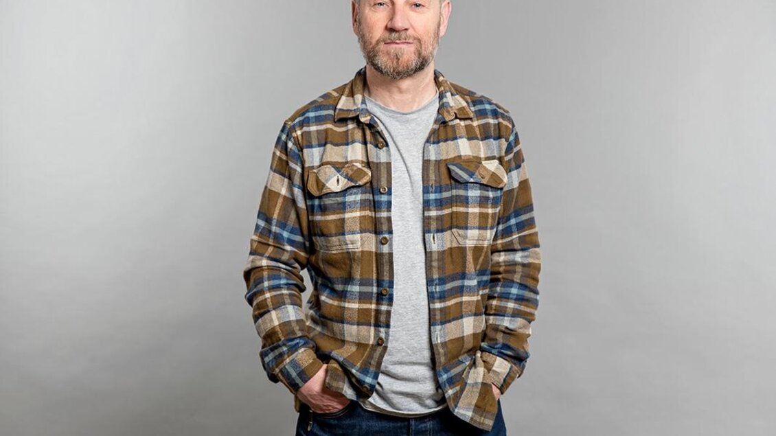 Ein Mann steht lächelnd vor einem grauen Hintergrund. Er trägt ein karierter Hemd und Jeans. | © ©Andrea Sojka