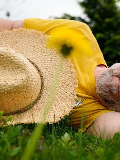 A man is relaxed lying on the grass, holding a straw hat in his hand. Around him, yellow flowers are blooming, and the atmosphere is calm and natural. | © Oststeiermark Tourismus