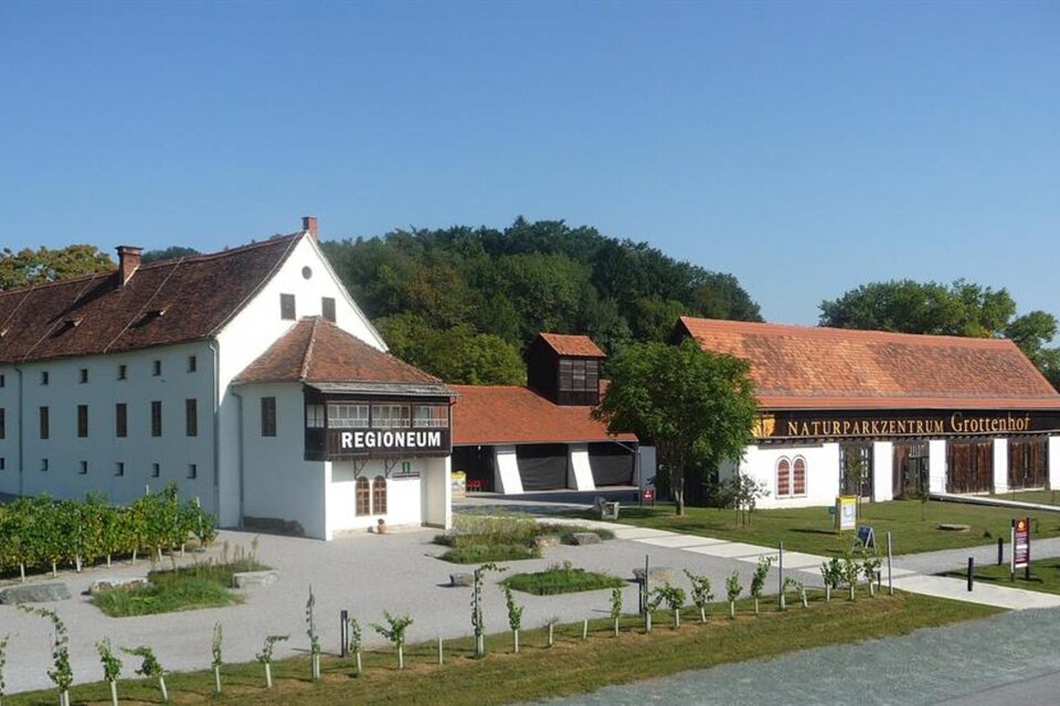 Ein historisches Gebäude mit weißen Wänden und roten Dächern steht unter klarem Himmel. Im Vordergrund ist ein gepflegter Garten zu sehen. | © Besucherzentrum Grottenhof