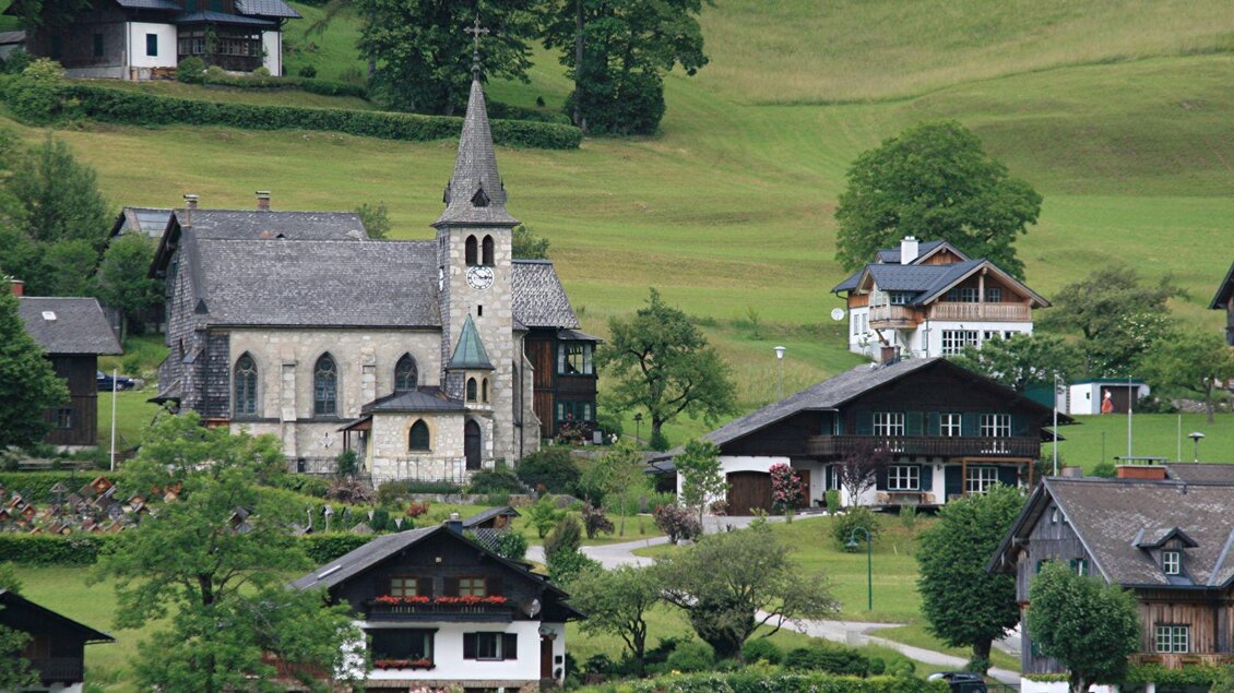 Ein malerisches Dorf mit einer kleinen Kirche und traditionellen Häusern. Die Umgebung ist grün und hügelig. | © Rabensteiner
