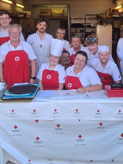 A group of people stands smiling in front of a table covered with a white cloth on which the logo of the Red Cross is visible. Most are wearing white clothing and red aprons. | © Rotes Kreuz Trofaiach
