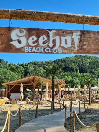 A sign of the Seehof Beach Club is in the foreground. In the background, beach loungers and a cozy wooden building can be seen. | © Oststeiermark Tourismus