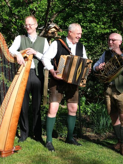 Three musicians are standing outdoors and playing traditional instruments. A harp, an accordion, and a euphonium can be seen. | © Helmut Fladenhofer