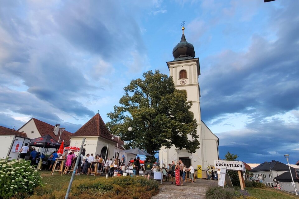 Eine neugotische Kirche mit einem hohen Turm steht im Mittelpunkt. Menschen sammeln sich in der Nähe und genießen eine Veranstaltung im Freien. | © TV Südsteiermark - Irene Löschnig