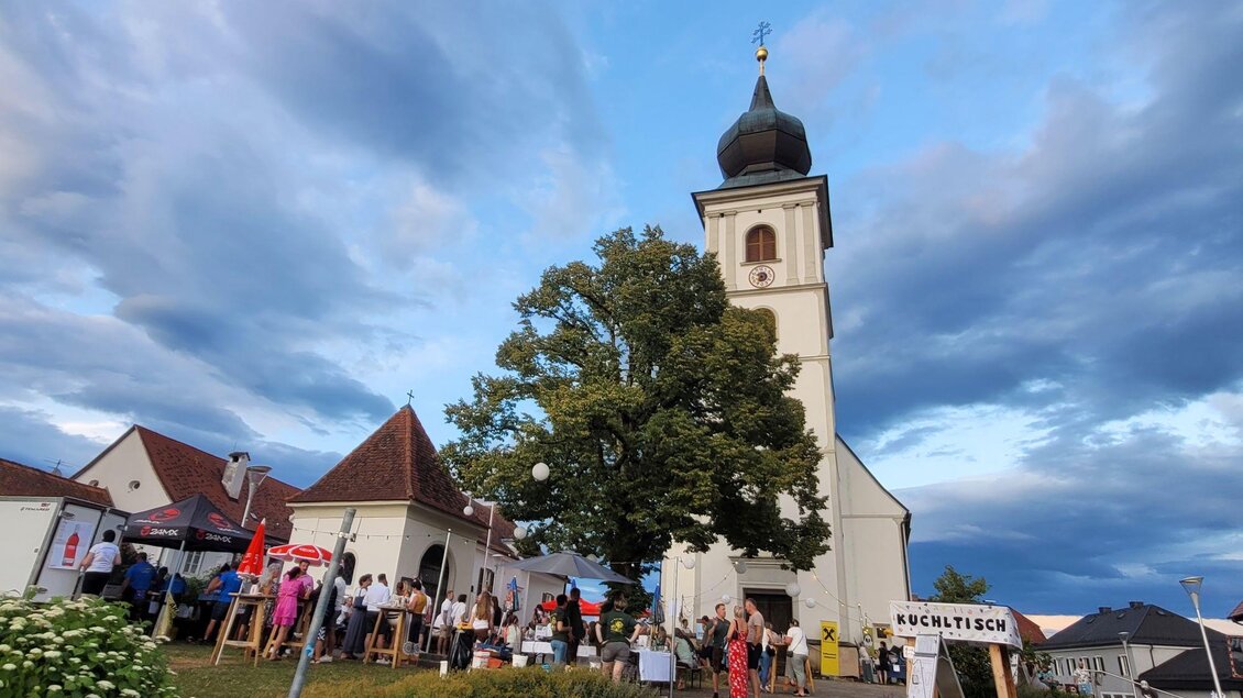 Eine neugotische Kirche mit einem hohen Turm steht im Mittelpunkt. Menschen sammeln sich in der Nähe und genießen eine Veranstaltung im Freien. | © TV Südsteiermark - Irene Löschnig
