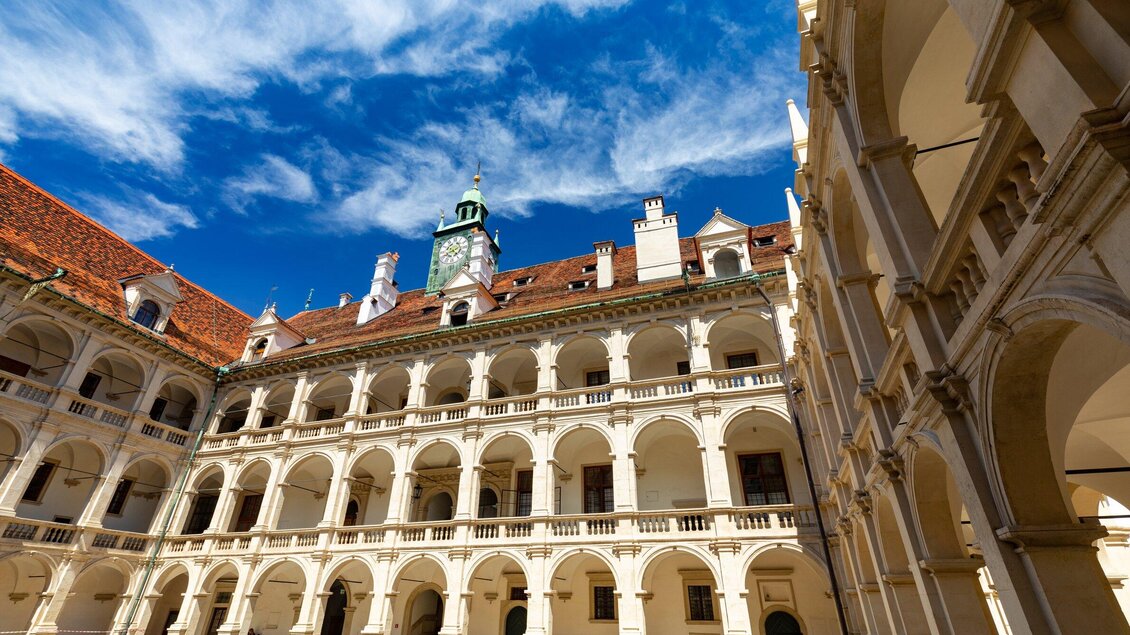 Ein schöner Innenhof mit historischen Arkaden und einem blauen Himmel. Im Hintergrund ist ein Turm mit einer Uhr zu sehen. | © Graz Tourismus - Harry Schiffer