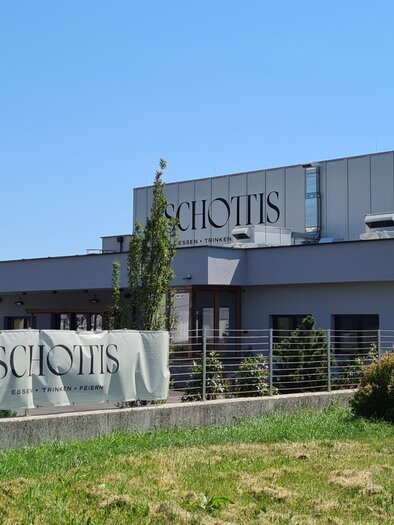 Ein modernes Gebäude mit der Aufschrift "Schottis" in einer grünen Umgebung. Die Sonne scheint klar am blauen Himmel. | © Christine Schwetz