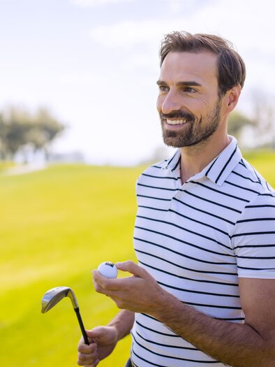 A smiling man stands on a golf course holding a golf club and a golf ball in his hand. In the background, lush green grass is visible. | © TV BW