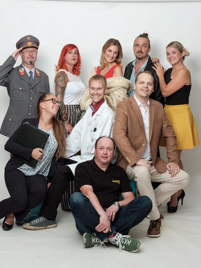 Eine Gruppe von Personen in verschiedenen Outfits posiert vor einem neutralen Hintergrund. Einige tragen historische Kleidung, während andere modern gekleidet sind. | © Theatergruppe Fladnitz