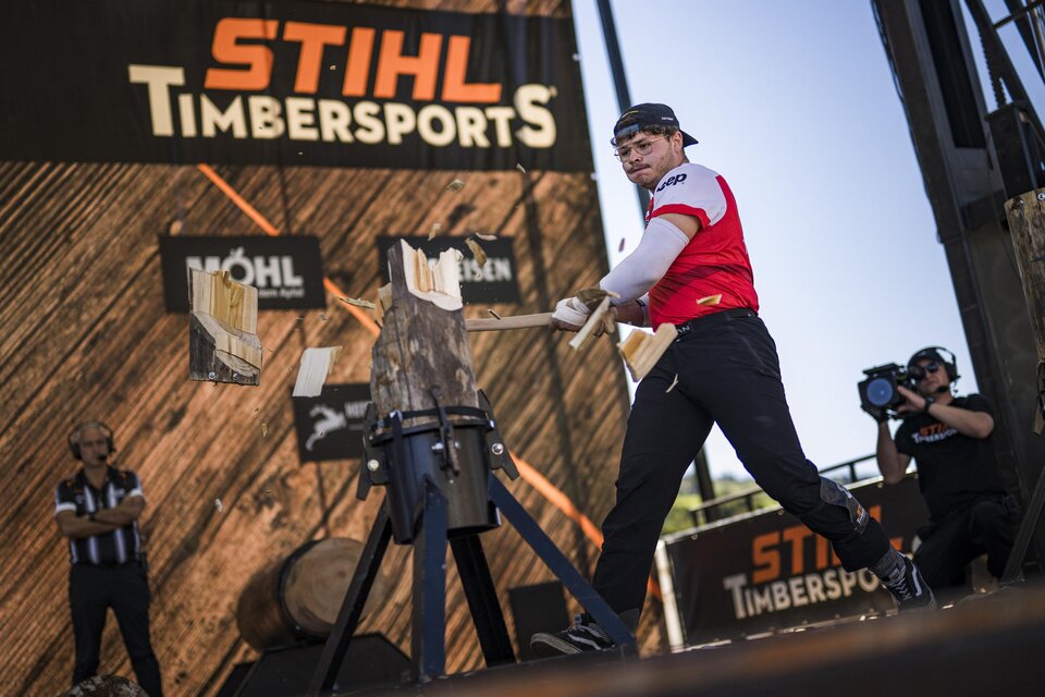 Ein Wettkämpfer bei den STIHL Timbersports schlägt auf ein Holzstück. Im Hintergrund sind Zuschauer und Sponsorenbanner zu sehen. | © STIHL