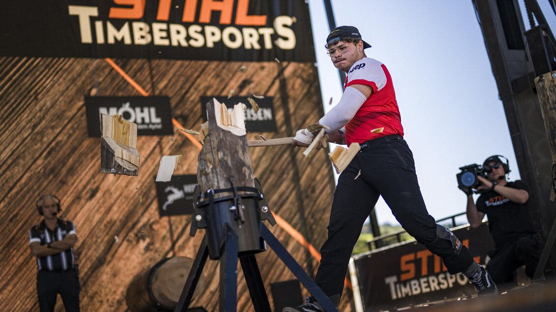 Ein Wettkämpfer bei den STIHL Timbersports schlägt auf ein Holzstück. Im Hintergrund sind Zuschauer und Sponsorenbanner zu sehen. | © STIHL