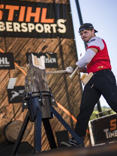 Ein Wettkämpfer bei den STIHL Timbersports schlägt auf ein Holzstück. Im Hintergrund sind Zuschauer und Sponsorenbanner zu sehen. | © STIHL
