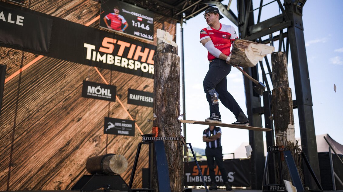 Ein Sportler präsentiert eine beeindruckende Leistung im Holzfäll-Wettbewerb auf einer Bühne. Im Hintergrund sind Sponsorenbanner und Holzstämme sichtbar. | © STIHL