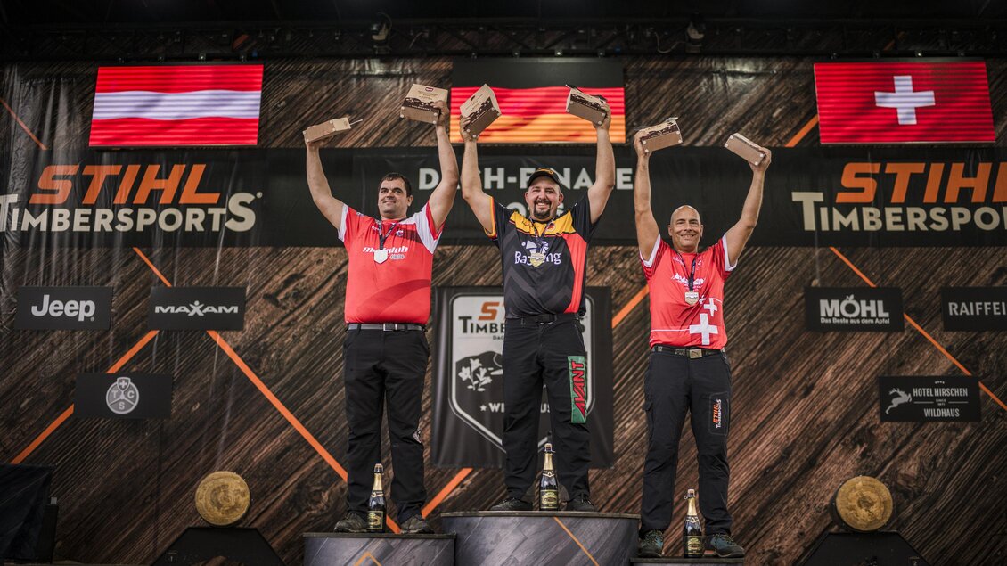 Drei Männer stehen auf Podesten und halten Trophäen in den Händen. Im Hintergrund sind Flaggen von Österreich, Deutschland und der Schweiz sowie das Logo von STIHL Timbersports zu sehen. | © Stihl