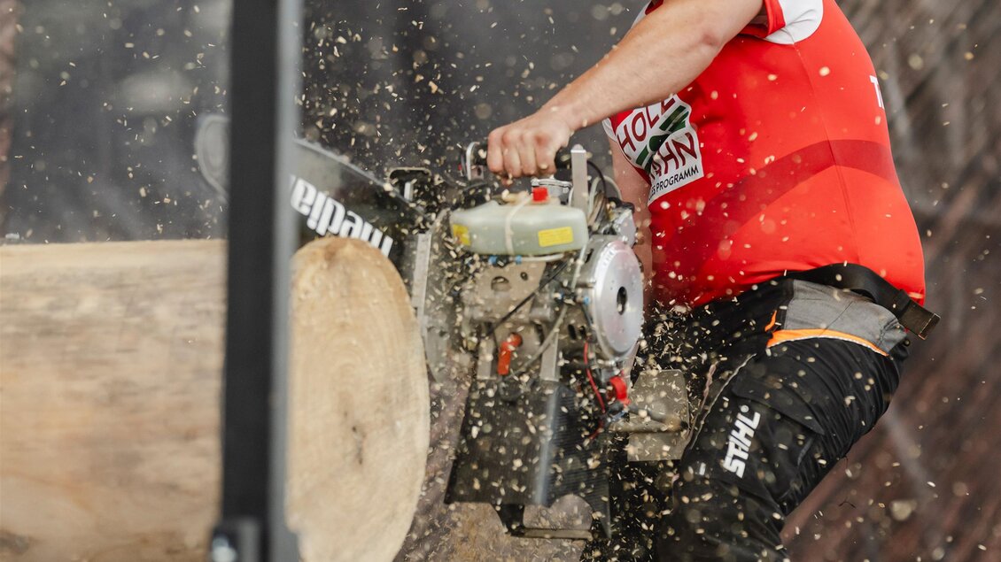 Ein Mann in einem roten Shirt arbeitet mit einer Kettensäge an einem Baumstamm. Holzspäne fliegen während des Schneidens in die Luft. | © Stihl