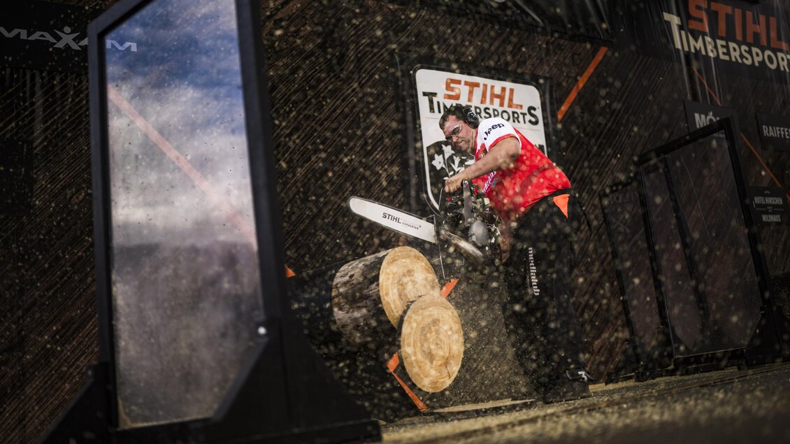 Ein Wettkämpfer schneidet mit einer Motorsäge einen Baumstamm. Im Hintergrund sind die Veranstaltungsbanner von STIHL Timbersports zu sehen. | © Stihl