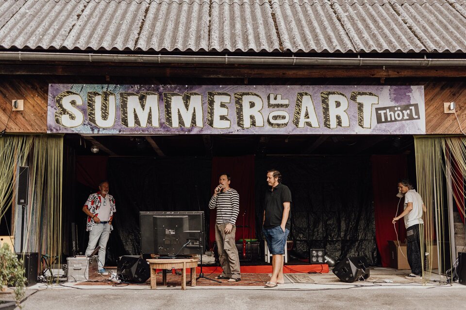 Eine Bühne mit einem Schild "SUMMER of ART Thörl" ist im Hintergrund sichtbar. Vier Männer stehen vorne und scheinen sich auf eine Veranstaltung vorzubereiten. | © nicoleseiser.at