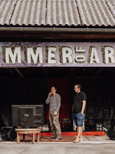 Eine Bühne mit einem Schild "SUMMER of ART Thörl" ist im Hintergrund sichtbar. Vier Männer stehen vorne und scheinen sich auf eine Veranstaltung vorzubereiten. | © nicoleseiser.at