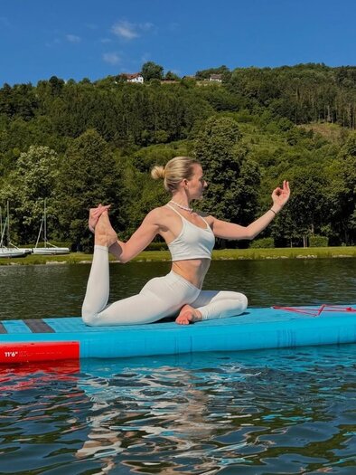 Eine Frau macht Yoga auf einem Stand-Up-Paddle-Board auf einem ruhigen Gewässer. Im Hintergrund sind Bäume und ein klarer blauer Himmel sichtbar. | © Yogina
