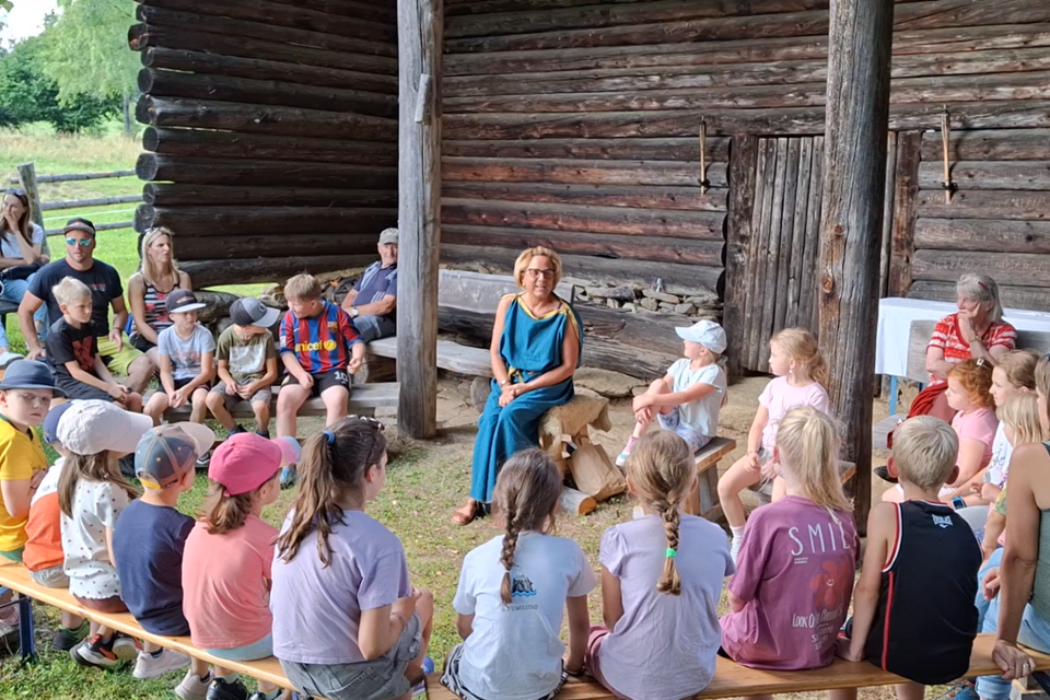 Eine Gruppe von Kindern und Erwachsenen sitzt in einem Freiluftraum vor einem Holzgebäude. Ein Erwachsener spricht in die Runde, während die Kinder aufmerksam zuhören.