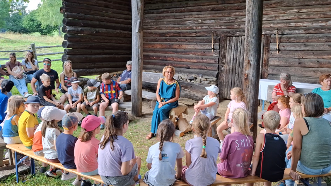 Eine Gruppe von Kindern und Erwachsenen sitzt in einem Freiluftraum vor einem Holzgebäude. Ein Erwachsener spricht in die Runde, während die Kinder aufmerksam zuhören.