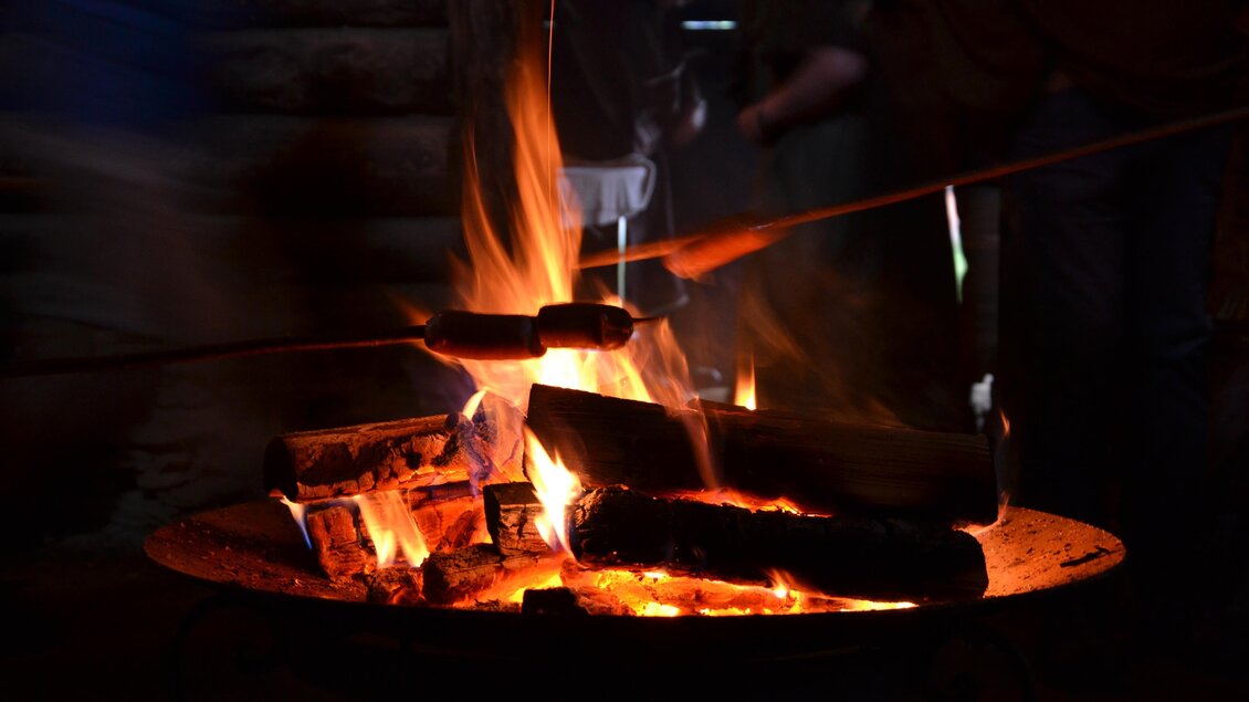 Ein gemütliches Lagerfeuer mit brennenden Holzscheiten und lodernden Flammen. Im Hintergrund sind Schatten von Personen zu sehen, die sich um das Feuer versammeln.