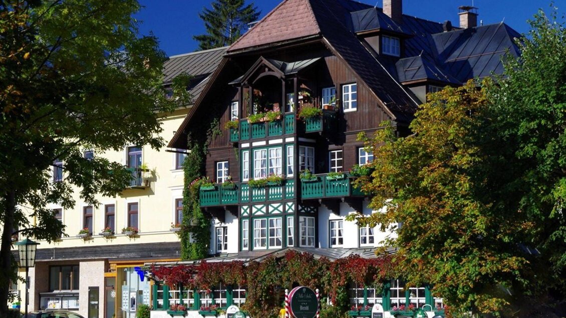 Eine sommerliche Außenansicht des Gasthof Berndl in Altaussee, ein traditionelles Ausseer Gasthaus mit Holzbalkonen und Blumen, umgeben von Bäumen und einem klaren blauen Himmel. | © Gasthof Berndl