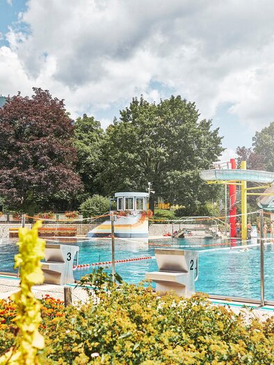 A swimming pool with a slide and surrounded by colorful flowers. The surroundings are green and sunny with clouds in the sky. | © TV Gesäuse