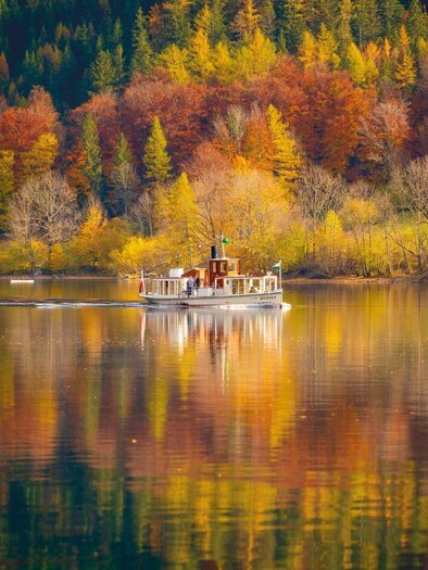 Ein nostalgisches Schiff fährt über einen ruhigen See, umgeben von bunten Bäumen im Herbst. Die leuchtenden Farben spiegeln sich im Wasser wider. | © Schifffahrt Grundlsee