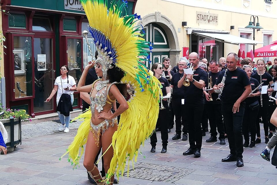 Eine Tänzerin in einem bunten Kostüm mit großen Federn präsentiert sich vor einer Menschenmenge. Im Hintergrund spielt eine Musikgruppe und es gibt festliche Stimmung. | © Tourismus Oststeiermark/Samba in Hartberg