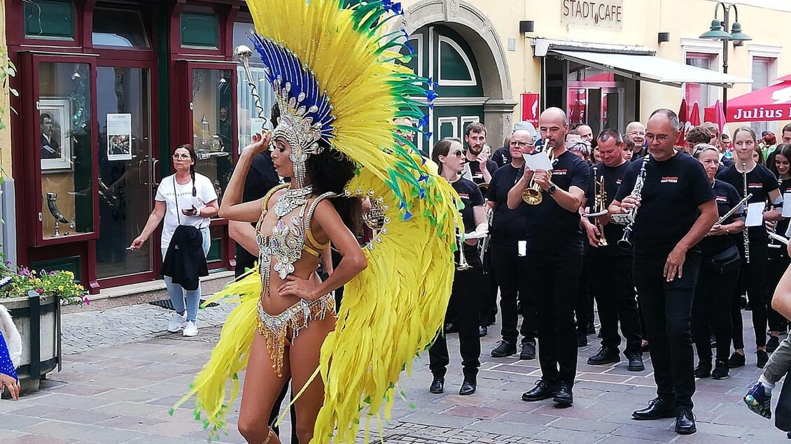 Eine Tänzerin in einem bunten Kostüm mit großen Federn präsentiert sich vor einer Menschenmenge. Im Hintergrund spielt eine Musikgruppe und es gibt festliche Stimmung. | © Tourismus Oststeiermark/Samba in Hartberg