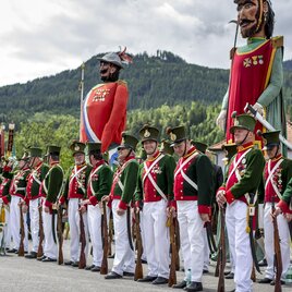 Ein festlicher Umzug mit Menschen in historischen Uniformen und großen Figuren. Im Hintergrund sind grüne Hügel und Wolken sichtbar. | © Steiermark Tourismus, ikarus.cc