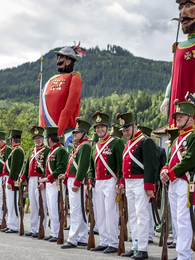 Ein festlicher Umzug mit Menschen in historischen Uniformen und großen Figuren. Im Hintergrund sind grüne Hügel und Wolken sichtbar. | © Steiermark Tourismus, ikarus.cc