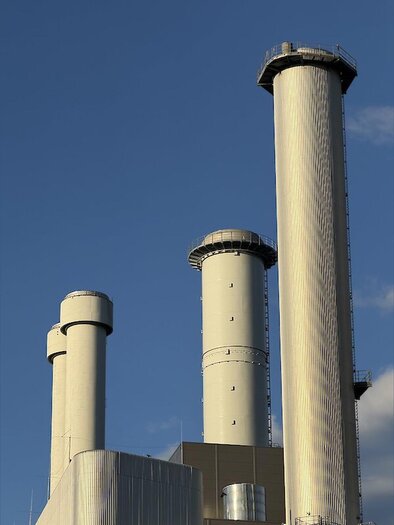 Drei hohe, zylindrische Schornsteine einer Industrieanlage mit Metallverkleidung und Wartungsplattformen vor blauem Himmel. | © Sascha Paar