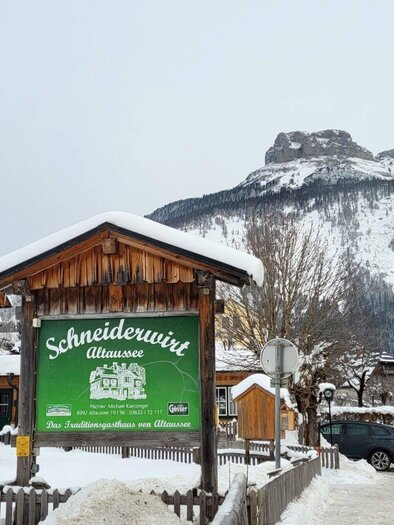 The Schneiderwirt tavern in the heart of Altaussee in wintry snow, surrounded by traditional wooden houses in "Ausseer" style. In the background, the majestic mountain Loser rises beneath a gray sky. | © Petra Kirchschlager