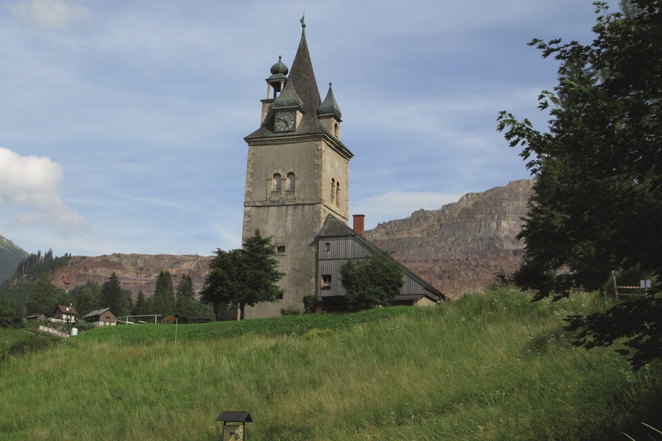 Ein historisches Gebäude mit dekorativen Fassadenmalereien und Fenstern. Im Hintergrund sind Berge zu sehen, bei klarem Himmel. | © Hermann Buder