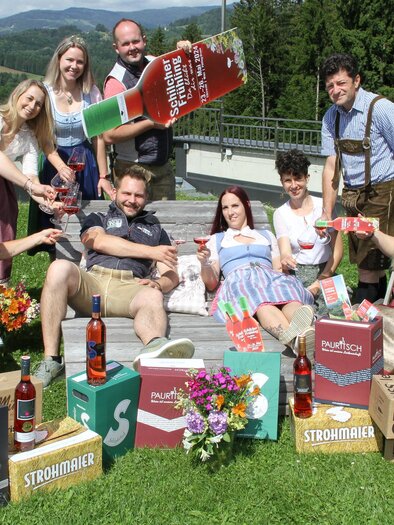 Eine fröhliche Gruppe von Menschen in traditionellen Trachten, die auf einer Wiese zusammen posieren. Sie halten Getränke und umgeben sich mit Kartons und Blumen. | © Josef Fürbass