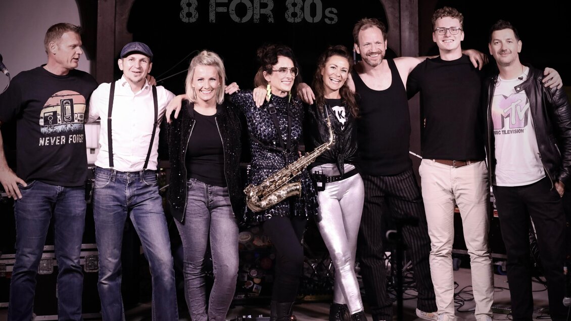 Eine Gruppe von acht Personen steht zusammen auf einer Bühne. Sie tragen verschiedene Outfits und lächeln in die Kamera. | © 7for80s