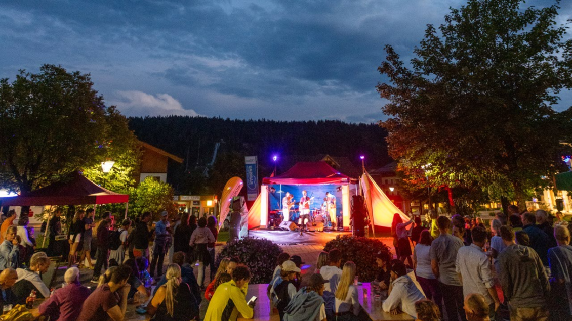 Ein Live-Konzert im Freien mit einer Menschenmenge. Die Bühne ist von bunten Lichtern und Bäumen umgeben, während die Atmosphäre festlich wirkt. | © Michael Simonlehner
