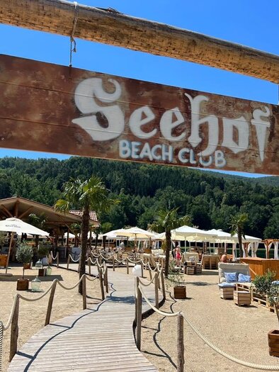 Ein einladender Strandclub mit einem Holzschild, das "Seehof Beach Club" anzeigt. Sonnenliegen und Schirme laden zum Entspannen ein, umgeben von einer schönen Naturlandschaft. | © Seehof-Beachclub