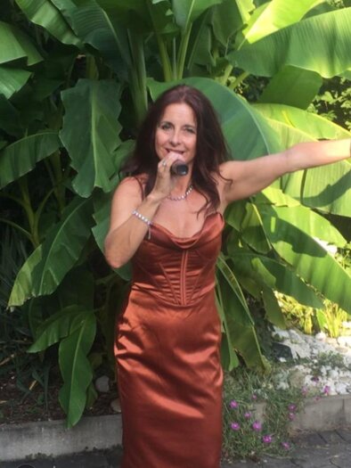 Eine Frau in einem eleganten, bronze-farbenen Kleid steht vor großen Pflanzen und hält ein Mikrofon in der Hand. Sie scheint zu singen oder zu performen. | © Christian Bernhard