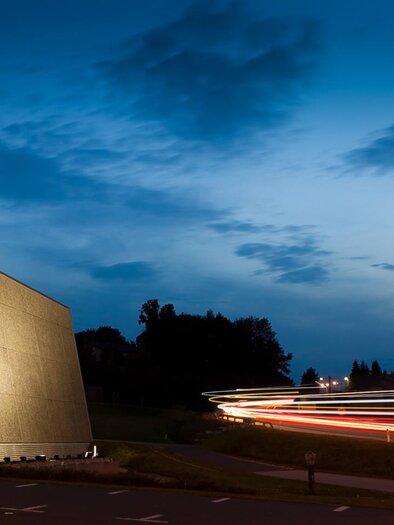 Ein modernes Gebäude mit dem Schriftzug "LANNACH" bei Dämmerung. Die Umgebung ist ruhig und es sind einige Lichtspuren von Fahrzeugen zu sehen. | © MG Lannach - Petru Rimovetz