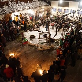Eine festliche Versammlung in einem beleuchteten Innenhof, umgeben von schneebedeckten Bäumen. Menschen stehen zusammen und halten Fackeln, während sie die festliche Stimmung genießen.