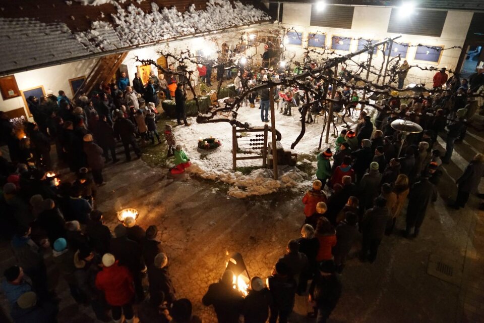 Eine festliche Versammlung in einem beleuchteten Innenhof, umgeben von schneebedeckten Bäumen. Menschen stehen zusammen und halten Fackeln, während sie die festliche Stimmung genießen.